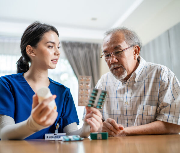 Asian Senior elderly male patient consult with physician nurse at nursing home care. Caregiver therapist pharmacist girl hold medicine pills bottle, explain prescription to older disabled guy in house