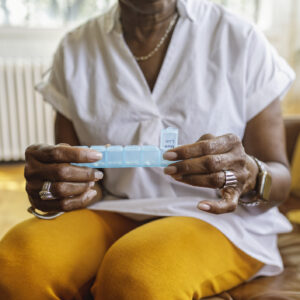 Senior black woman sits on the couch at home and takes medications from a daily pill organizer. Cropped shot does not show the woman