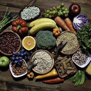 Dietary and healthy food themes. Table top view of fresh vegetables and legumes on rustic wooden table. Food is rich of fiber ideal for dieting and healthy eating. Includes corn, avocado, broccoli, orange fruit, grapes, bell pepper, lettuce, banana, apple almonds and wholegrain pasta.
